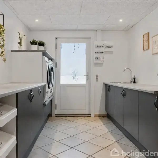 Vaskemaskine og tørretumbler på række i et funktionelt vaskerum med mørkegrå skabe, hvide vægge, fliser på gulvet og en glasdør ud til en vinterhave. Pflanzen på hylder tilføjer liv.