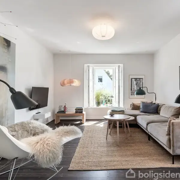En stue med moderne møbler som en hvid lænestol med pelsbeklædning, grå sofa, og runde træborde. Rummets vægge er dekoreret med kunst, og store vinduer lader lys komme ind.