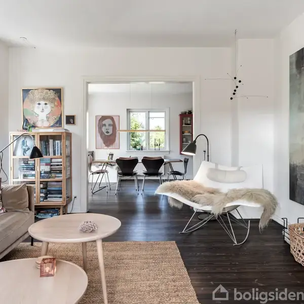 Sofa og runde borde står i en moderne stue med trægulv, store vinduer og kunst på væggene. Et værelse med spisebord ses i baggrunden.