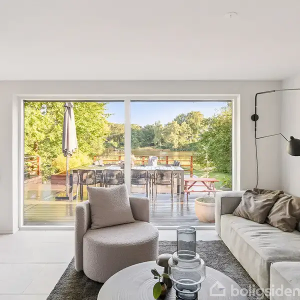 En stue med en grå stol og sofa foran store glasdøre. Udenfor er en træterrasse med spisebord og parasol omgivet af frodige grønne træer ved en sø.