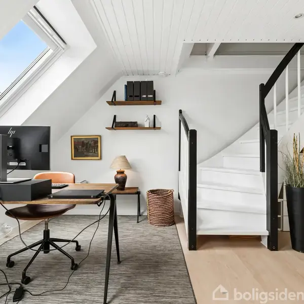 Et moderne kontor med et skrivebord og computer; en trappe fører opad. Rummet har skråvægge med ovenlysvindue, en lys indretning med planter og hylder.