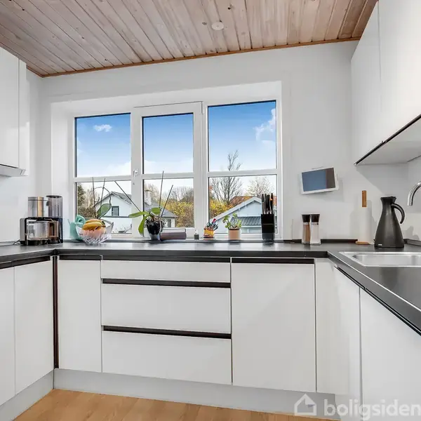 Et moderne køkken set fra hjørnet med hvide skabe og en sort bordplade. Vinduer viser en udsigt til en klar blå himmel og træer.