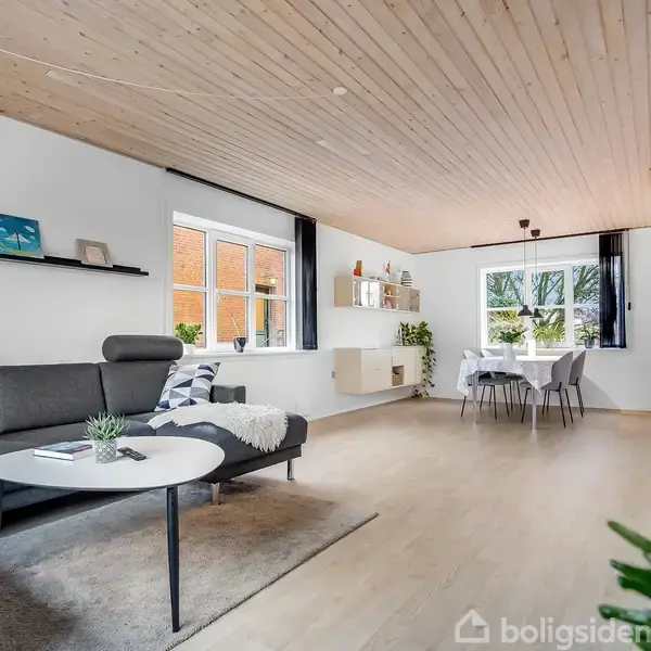 Stue med sort sofa placeret ved et rundt sofabord. Rummet har lyse trævægge og lofter, store vinduer, og et spisebord med stole i den anden ende.