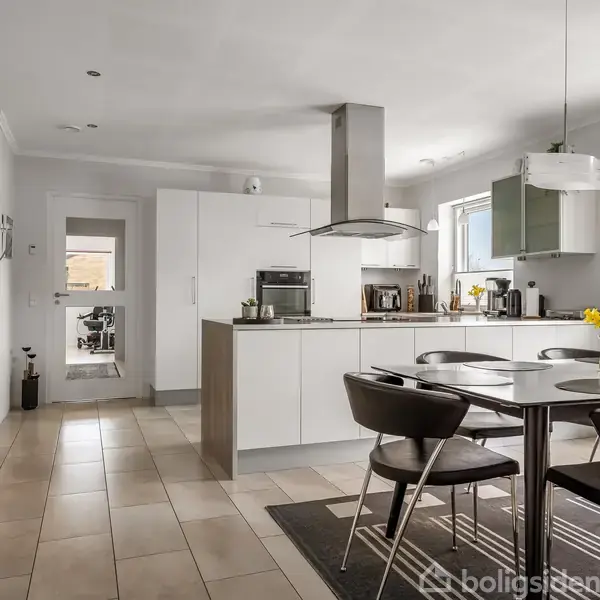 Køkken med moderne, hvidt interiør, indeholder et spisebord med fire stole, blomster på bordet. Åben layout med vinduer, lysindfald og en ø i midten.