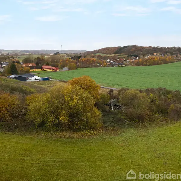 Grønne marker med træer i efterårsfarver omkranser et landsbyområde med røde tage, placeret i en bakket landskab under en klar blå himmel.