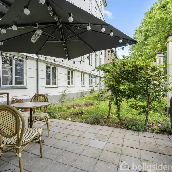 Terrasse med bord og stole står under en stor parasol med lyskæder. Omgivet af en hvid bygning og frodig have med træer, viser en hyggelig udendørs opholdssted.