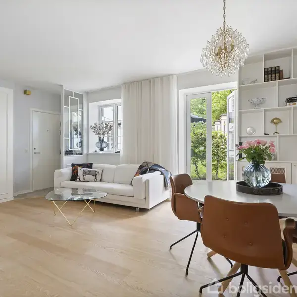 En lys stue med en hvid sofa til venstre, en rund spisebord med fire brune stole til højre og en reol fyldt med dekorative genstande i baggrunden. Store vinduer lukker lys ind.