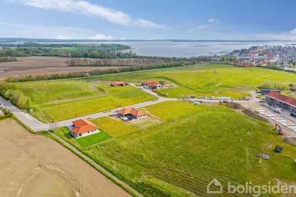 Flere nybyggede huse med røde tage er placeret i et grønt landskab. Området er åbent med udsigt til en nærliggende by og vand i baggrunden.
