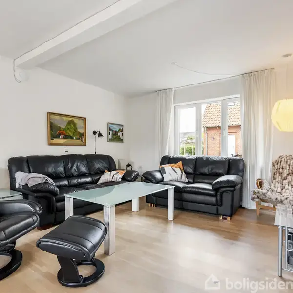 Stuen har sorte lædermøbler, herunder en sofa og lænestol, samt et glasbord. Rummet er lyst med et vindue til venstre og dekorative malerier på væggene.