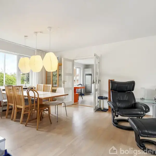 En moderne stue med et spisebord i træ og seks stole. Tre pendellamper hænger over bordet. Til højre er der en sort læderlænestol og sofa. Store vinduer lukker dagslys ind.