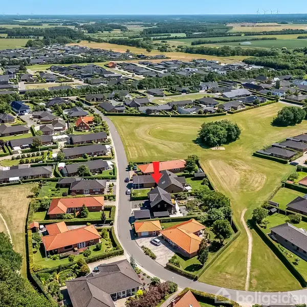 Oppefra ses et villakvarter med huse omgivet af grønne områder. En rød pil peger på et bestemt hus midt i kvarteret. Landskabet omkring er fladt og grønt.
