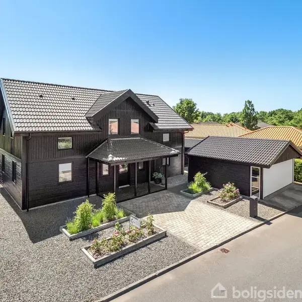 Sort træhus står stille ved en asfalteret vej, omgivet af en grusbelagt have med grønne buske. Ved siden af huset er en mindre garage.