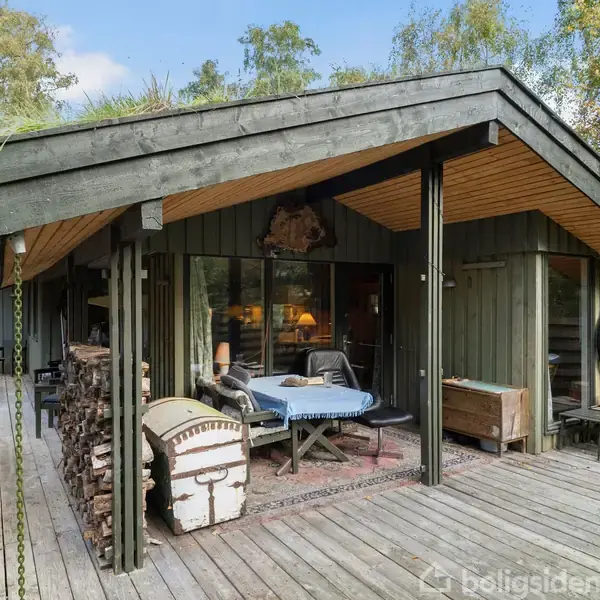 Et træhus med en rummelig overdækket terrasse, omgivet af skovbevoksning. På terrassen er der en pejs, brændestabel og et bord med stole, hvilket skaber en hyggelig atmosfære i naturen.