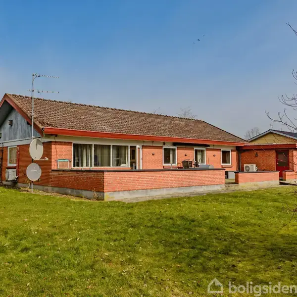 Rødt murmestervilla står stille på en grøn græsplæne under en klar blå himmel. Omkringliggende huse og en trægrænse danner baggrund. To paraboler ses på husets side.