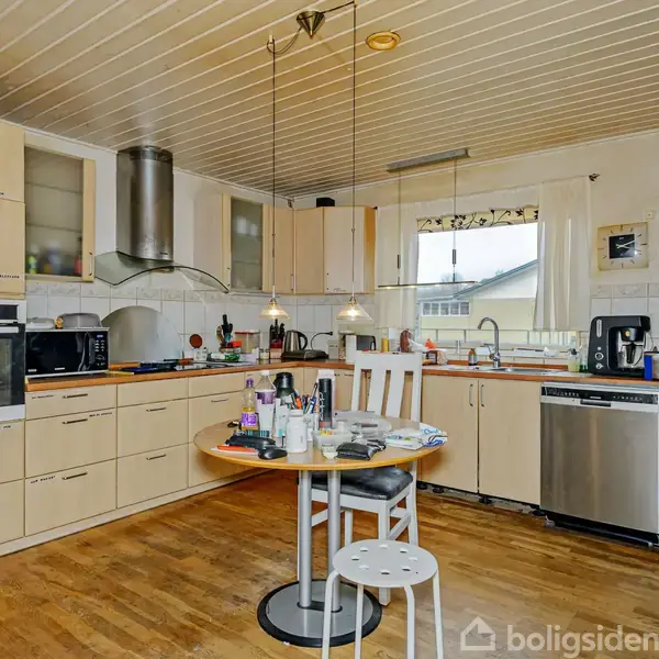 Køkken med lyse træskabe og stålapparater, herunder ovn og stort køleskab. Rundt bord med stole står midt i rummet. Trægulv og hvidt loft skaber en lys og venlig atmosfære.
