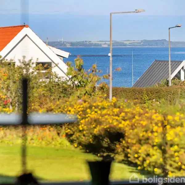 Et hus med rødt tag ses gennem et vindue, omgivet af en have med blomster. I baggrunden er der udsigt til hav og landskab i solskin.