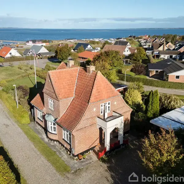 En rød murstensvilla med rødt tegltag står på en villavej. Actions: Ingen aktivitet. Context: Haver omgiver huset, og der er udsigt til havet i baggrunden.