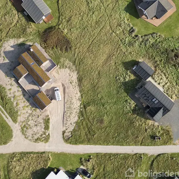 Et hus ligger midt i et græsområde med flere andre huse rundt omkring. En grusvej snor sig forbi grunden. En bil er parkeret ved huset.