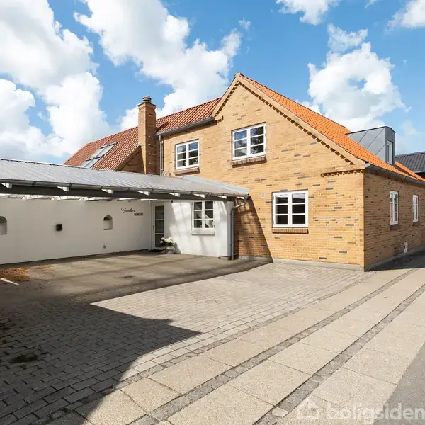 Et murstenshus med rødt tegltag står ved siden af en tom carport. Foran huset er en bred flisebelagt indkørsel. Det er solrigt, med skyer på himlen.