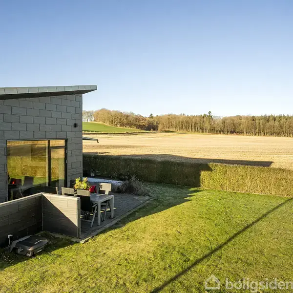 Et moderne hus med fladt tag står ved siden af en terrasse med havemøbler. Baggrunden består af en åben mark med skove i horisonten under en blå himmel.