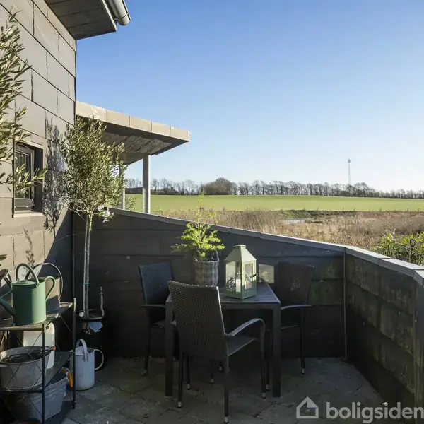 En terrasse med et bord og fire stole omgivet af grønne planter. Bordet står stille, mens landskabet i baggrunden viser åbne marker under en klar, blå himmel.