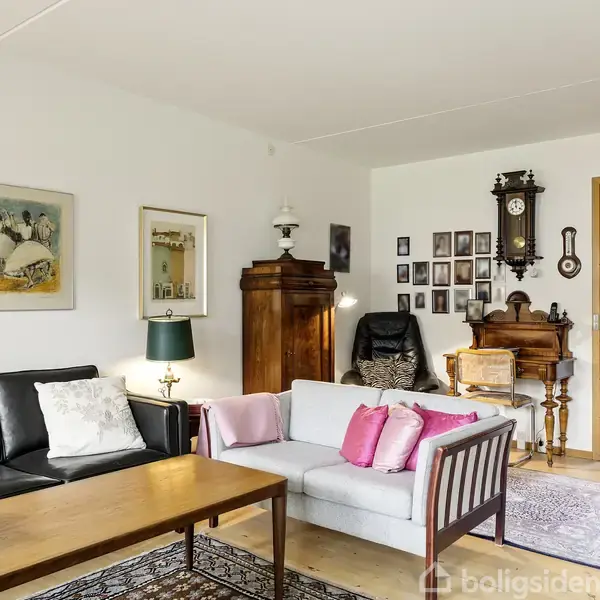 Sofaer står i stue med malerier på væggene. Farverige puder er placeret på den hvide sofa. Et klaver og en antik pendulur er baggrundsdetaljer, omgivet af tæpper og træmøbler.