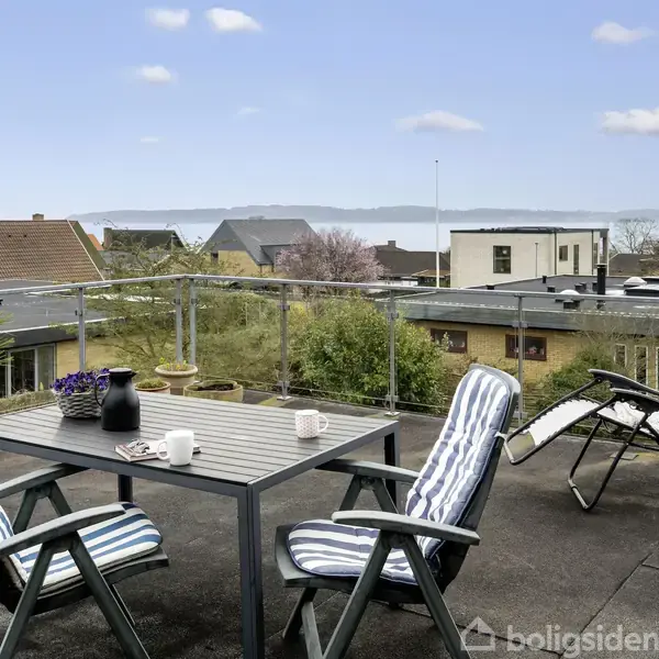 Et terrassebord med to stole og en liggestol står på en tagterrasse. Der er udsigt over huse med træer og havet i baggrunden. Himlen er blå med få skyer.