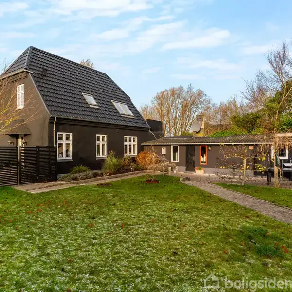 Et sortmalet hus med gråtag, omgivet af en stor have med buske og træer, står i et åbent område. En flisebelagt sti strækker sig mod en terrasse ved siden af huset.