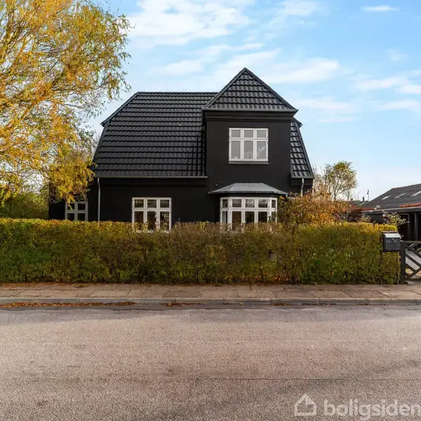 Et sort hus med gavl, omgivet af en hæk og træer, står ved en vej under en blå, skyfri himmel.