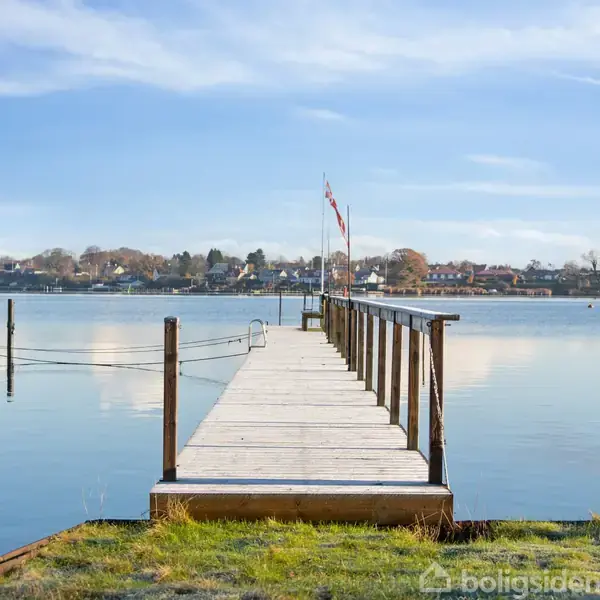 En træbro strækker sig ud i en stille sø, hvor flag vajer i vinden. I baggrunden ses en by med huse langs kysten under en blå himmel.