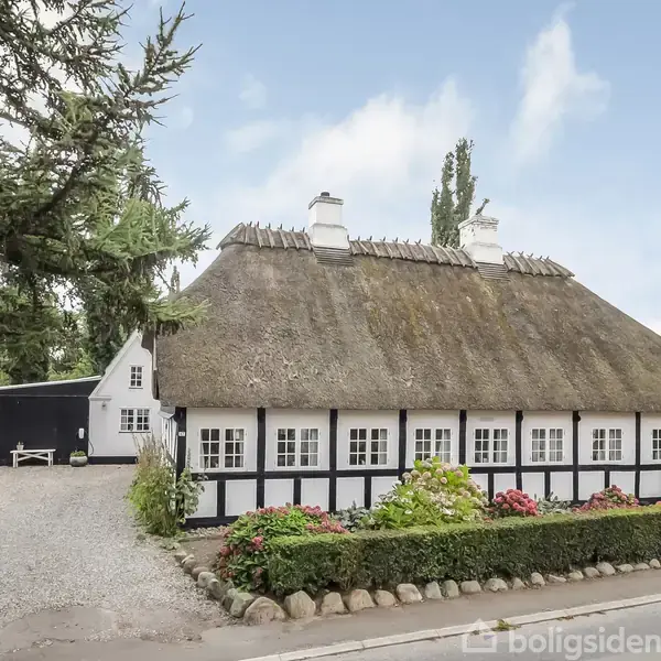 Bindingsværkshus med stråtag står stille. Detaljeret facade med hvide vinduer og sort struktur. Omkranset af have med blomsterbede, beliggende ved en grusvej med blå himmel i baggrunden.
