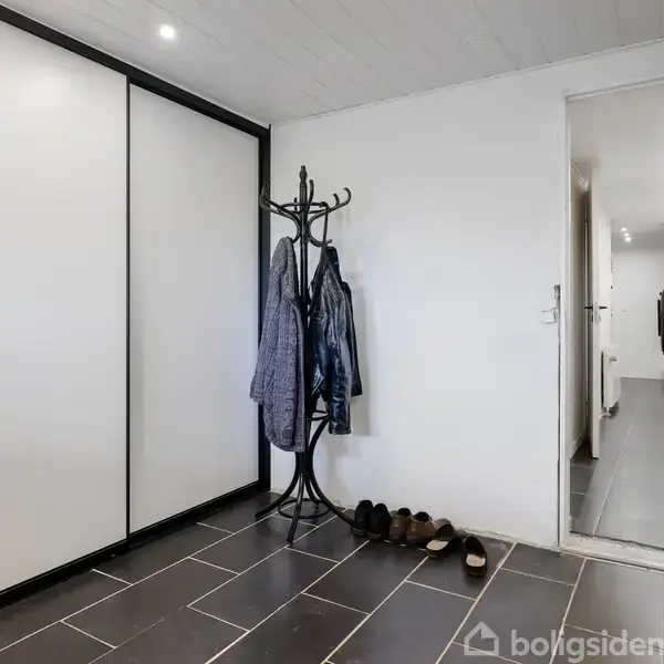Stumtjener med jakker står i entre med mørkt klinkegulv, mens flere sko er placeret ved siden af. Indbyggede skabe ses til venstre, og en dør fører videre ind i huset.