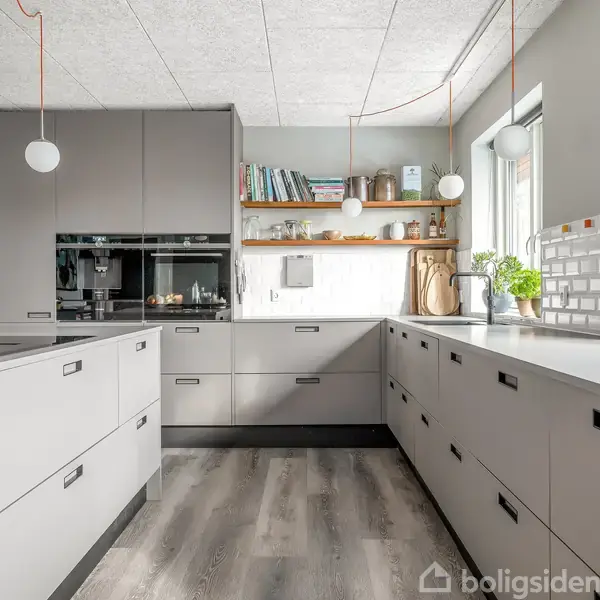 Gråt køkken med hvide lofthængte lamper. Køkkenet har indbyggede apparater, åbne hylder med bøger og planter samt vinduer, der giver naturligt lys. Gulvet er i trælignende materiale.