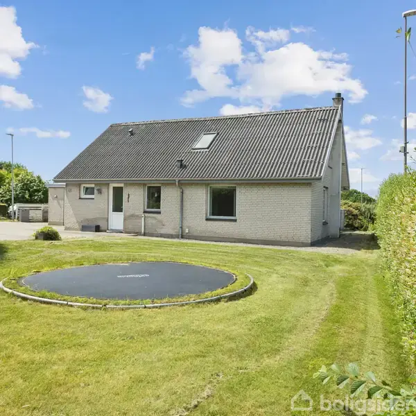Et hvidt hus med skråt tag står i en solbeskinnet have. En nedgravet trampolin er synlig i græsplænen foran huset, omgivet af grønne hække og en klar blå himmel.