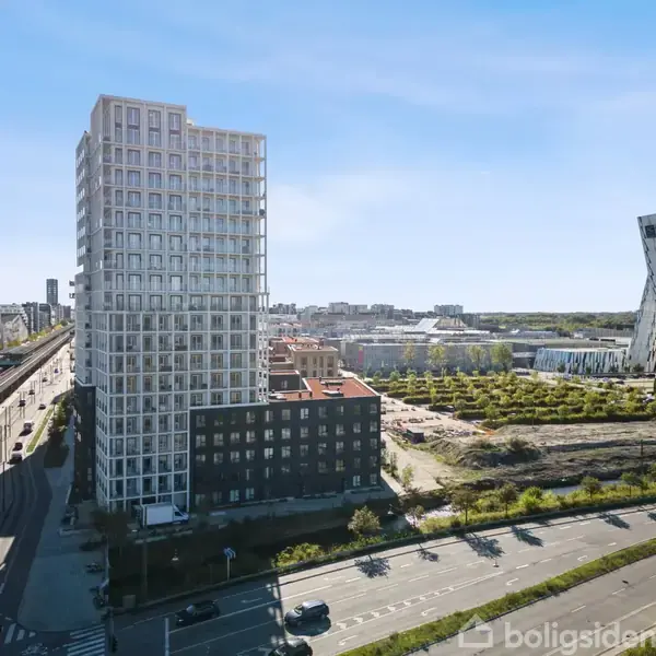 Et højt, moderne lejlighedsbyggeri står langs en bred vej, som har let trafik. I baggrunden ses flere bygninger og et åbent grønt område under en klar blå himmel.