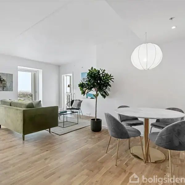 Rundt hvidt spisebord med fire grå stole står i en moderne stue. I baggrunden ses en grøn sofa, stort vindue og en plante, alt på trægulv.