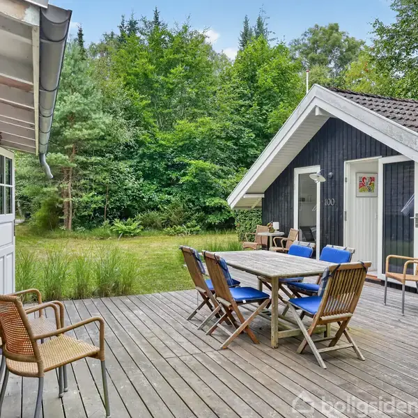 Sort træhus med opført træterrasse; bord og stole står rundt om bordet. Tæt skov i baggrunden giver en naturskøn atmosfære. Terrasse skaber overgang mellem hus og natur.