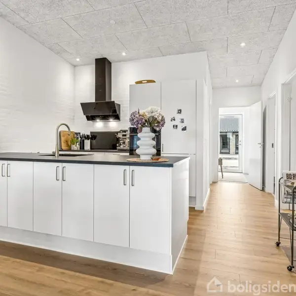 Køkken med hvidt skab og sort bordplade, blomster i vase og stålvaske. Baggrund viser en lys gang med trægulv og døråbninger.