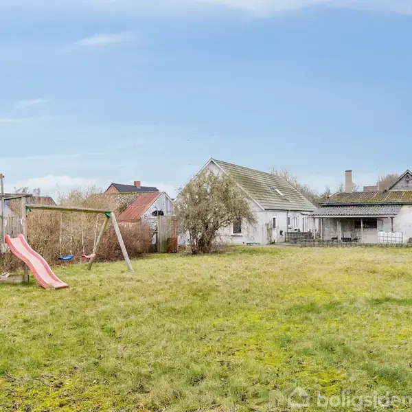 En gyngestativ med en rutsjebane står på en stor, græsbeklædt have. I baggrunden ses et hvidt hus med en terrasse og flere omkringliggende boliger.
