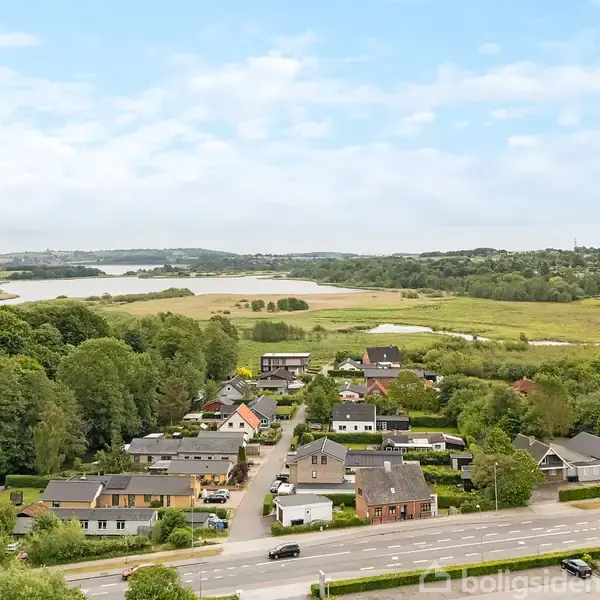 Boliger strækker sig langs en vej med skove til venstre, åbne marker og en sø i baggrunden. Landskabet har en blanding af bebyggelse og natur, som skaber en rolig atmosfære.