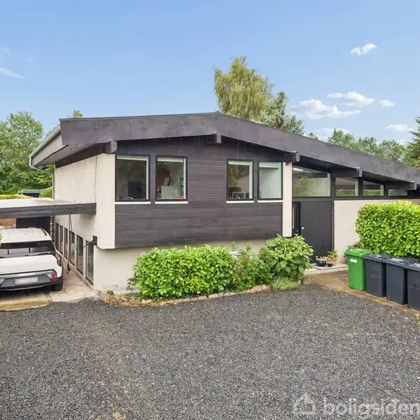 Hus med mørkt træværk, grå sokkel, står foran en grusbelagt indkørsel med affaldscontainere. To biler er delvist synlige under en carport. Omgivet af grønne buske og træer.
