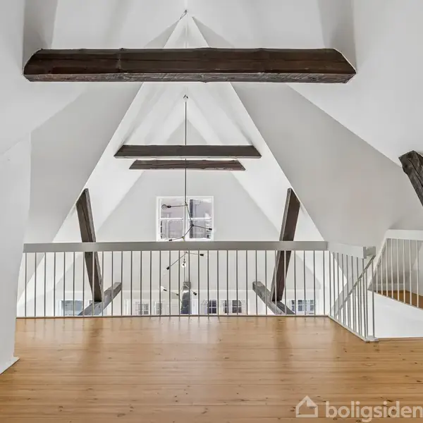 Loft med synlige træbjælker, lyst trægulv og hvidmalede vægge danner rammen om et rummeligt, åbent rum. En stor hængende lampe ses nedenfor i det underliggende værelse.