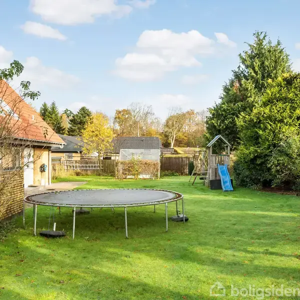 En trampolin står på en grøn græsplæne i en have omgivet af træer og en rød flagstang. Et hus med rødt tag ses til venstre, og en legeplads er bagved.