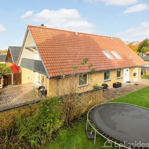 Hus med rødt tegltag, hvide vinduer, og terrasse på bagsiden, stående i en velplejet have med stor trampolin. Omgivet af andre huse og træer i efterårsfarver.