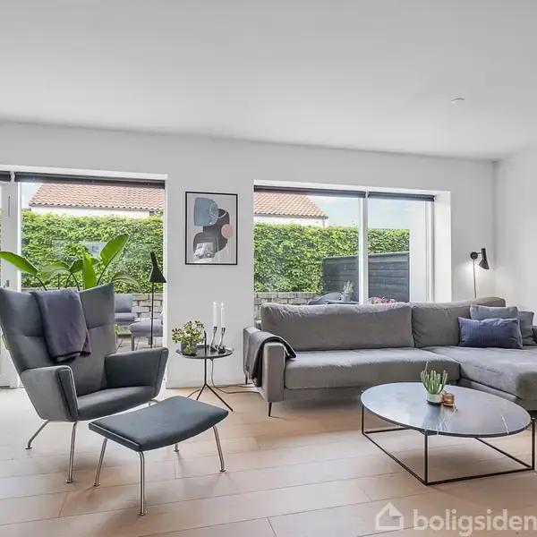 En moderne stue med en grå hjørnesofa, et sofabord og en lænestol foran store vinduer. Udenfor ses en grøn hæk og en terrasse med havemøbler.