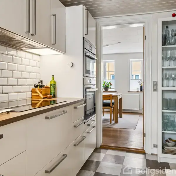 Et moderne køkken med hvide skabe og træbordplade. Skuffer trukket ud. Glasdør afslører glas og kopper. I baggrunden ses en spisestue med stort vindue og planter.