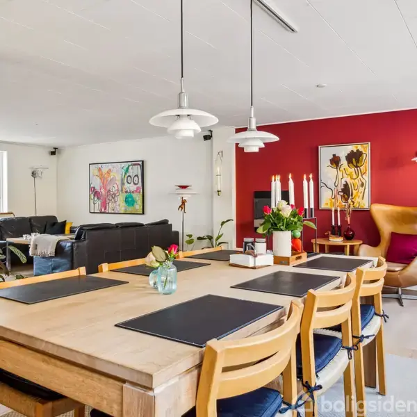 Spisebord med seks stole står i en spisestue med lyse træmøbler. I baggrunden ses en rød væg med kunstværker og en lænestol, omgivet af grønne planter og stearinlys.