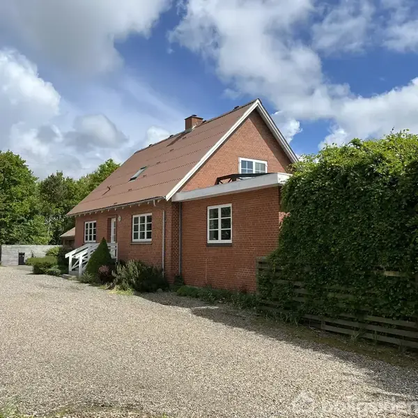 Rødt murstenshus med spidst tag, omgivet af træer og buske, står på en grusbelagt gårdsplads. Skyerne dækker delvist en blå himmel.