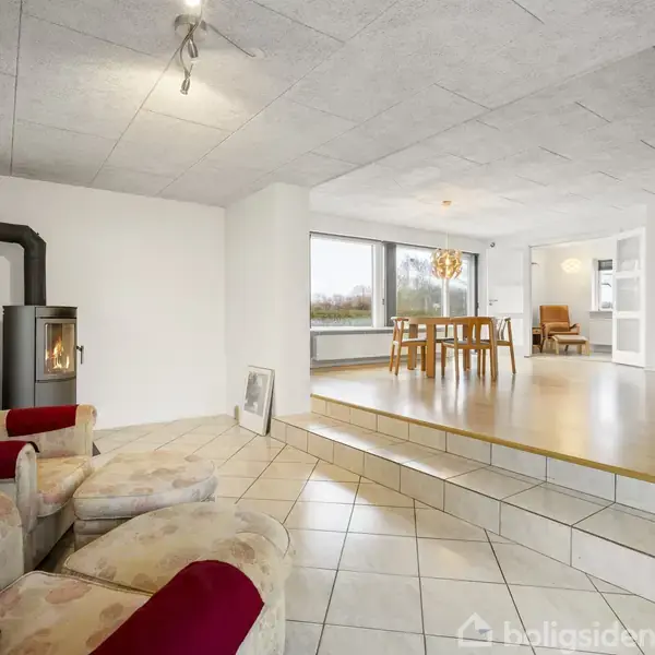 Pejs med ild i hjørnet, står i en lys stue med trin op til en spiseplads med træstole og bord, omgivet af store vinduer og lyse gulve.