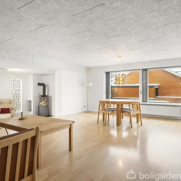 Stue med lyse trægulve og hvidmalede vægge, hvor en sofagruppe står ved væggen. En brændeovn brænder i hjørnet, og et spisebord er placeret tæt på store vinduer.